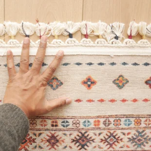 Illustration - Ihr Experte für schönes Wohnen - Eine Nahaufnahme einer Hand, die auf einem wunderschönen, farbenfrohen Vintage-Teppich aus der Vergangenheit platziert ist.